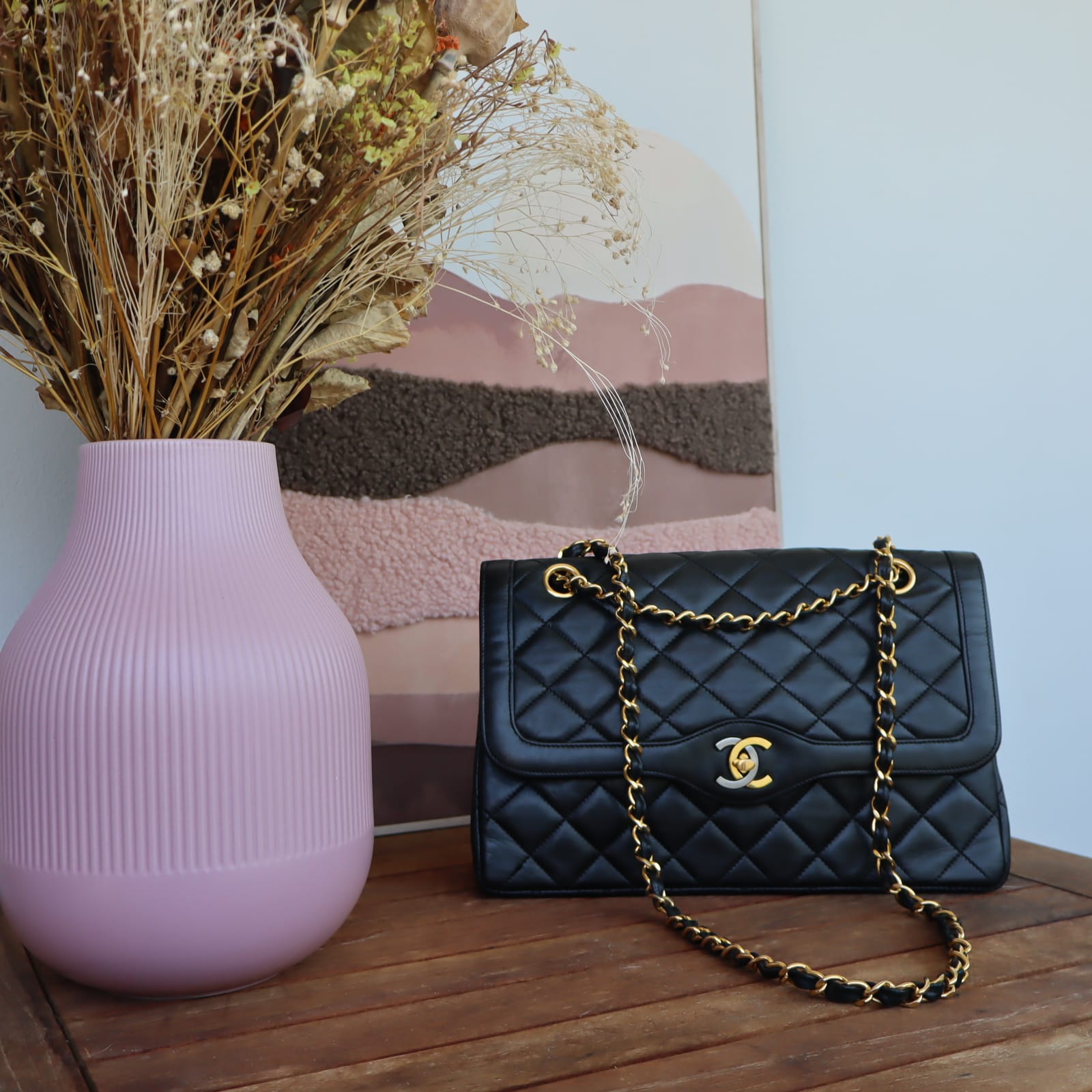 Sac Chanel Double Rabat en cuir noir matelassé, chaîne dorée entrelacée de cuir, posé à coté d'un vase.